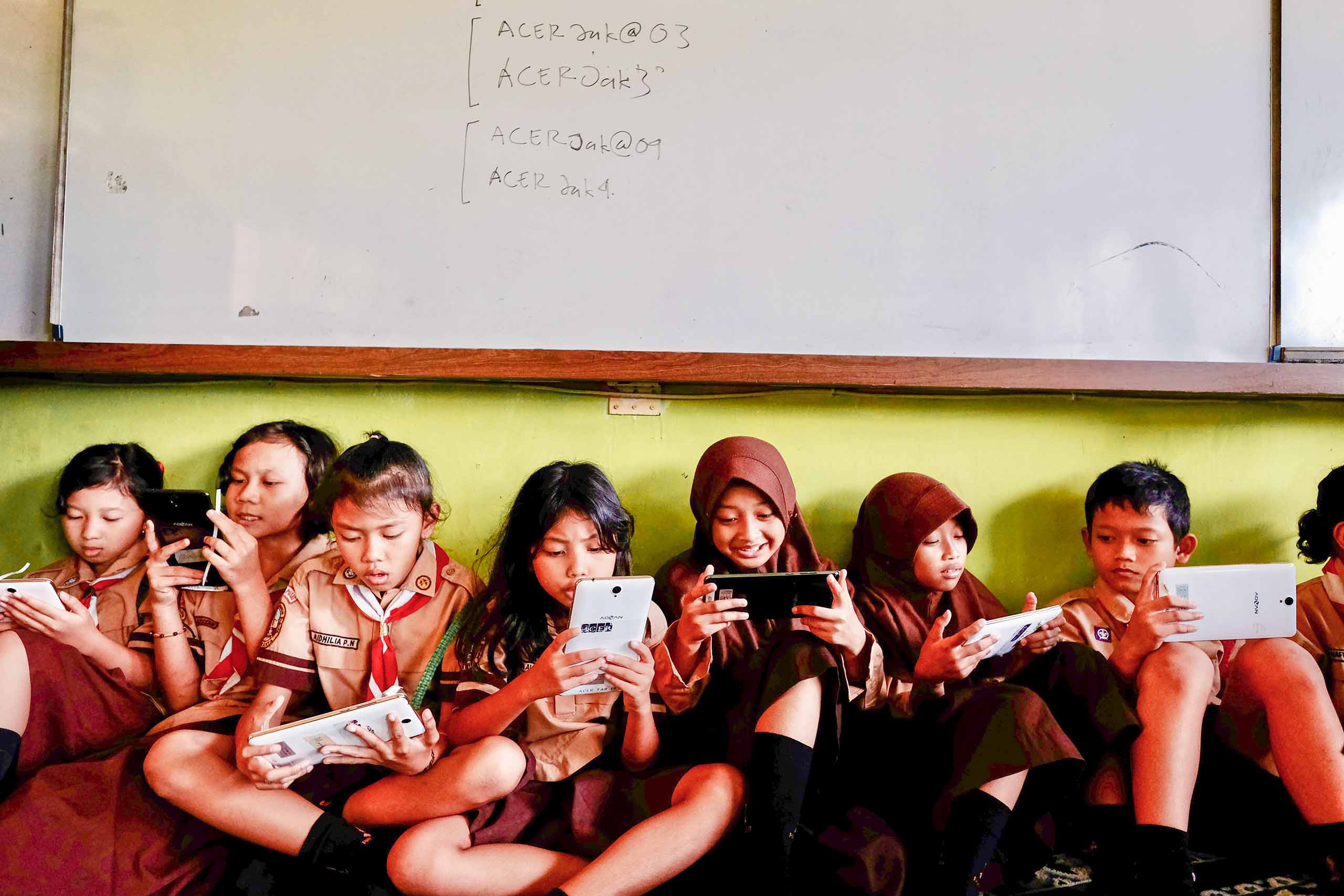 Children in school uniform sat with their backs to a wall using tablets and phones