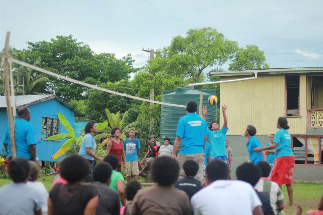 Photo of 6 people wearing matching blue t-shirts playing volleyball being watched by a group of people